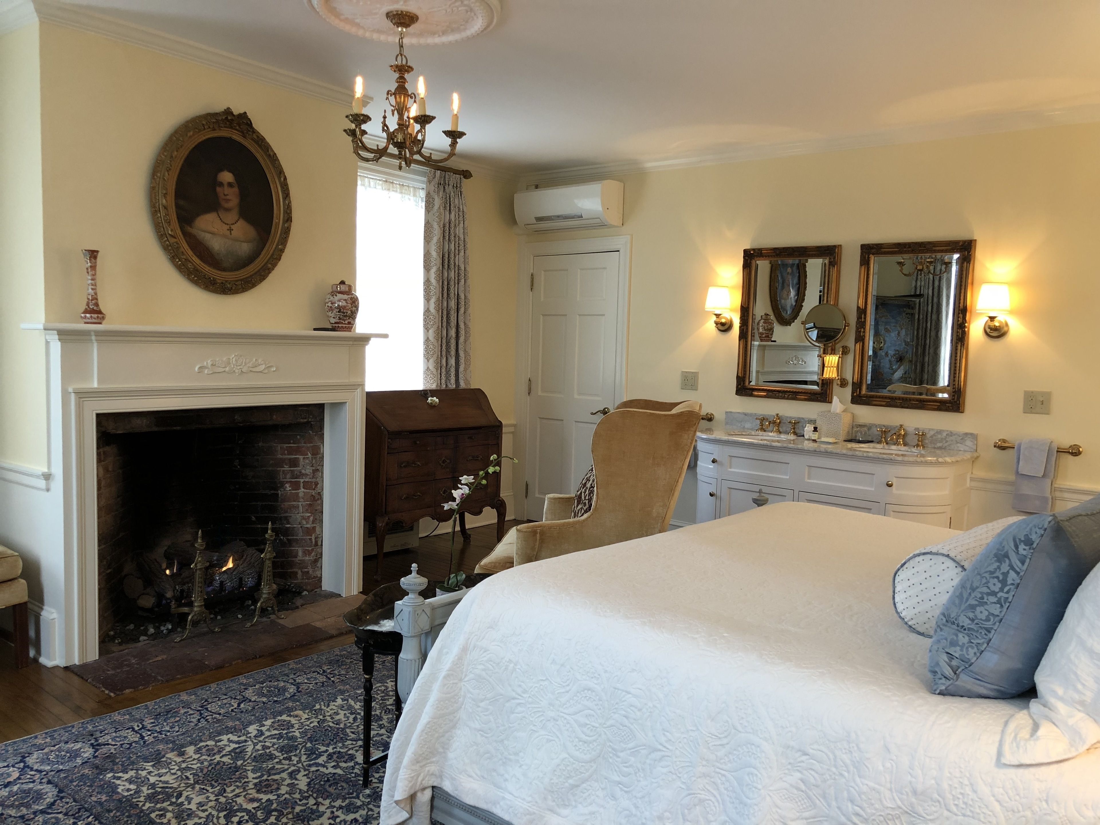 A bedroom with a large bed, a fireplace, a chandelier, a mirror, and a dresser.
