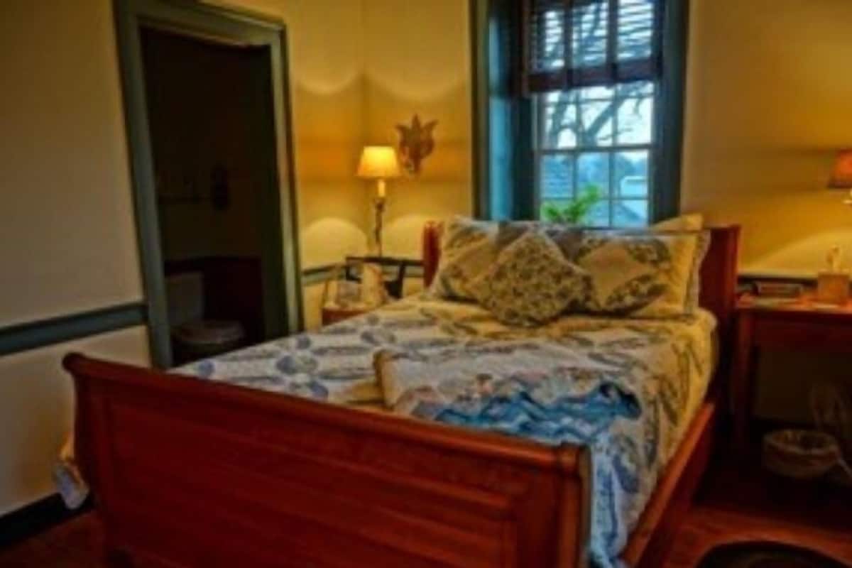 A wooden bed with a patterned bedspread, a nightstand with a lamp, and a window with a view of the outside.