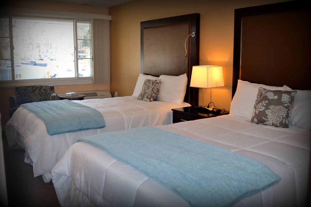 A hotel room with two beds, a window with a view, a bedside table with a lamp, and a decorative pillow.