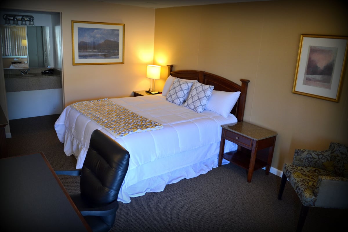 A hotel room with a bed, bedside table, lamp, chair, desk, and a small table with a lamp.