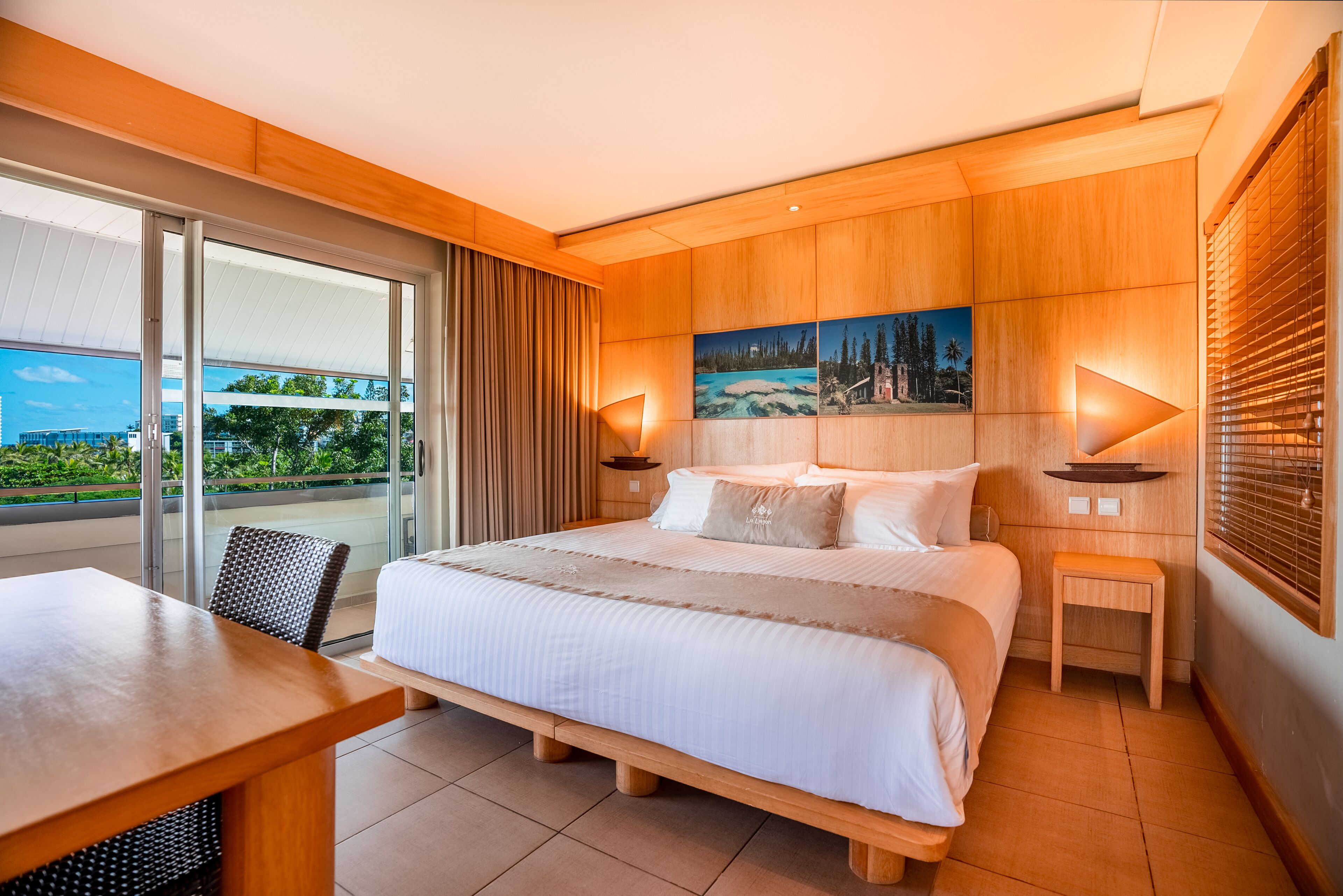 A hotel room with a large bed, a wooden headboard, a dining table, and a view of the outdoors through a sliding door.