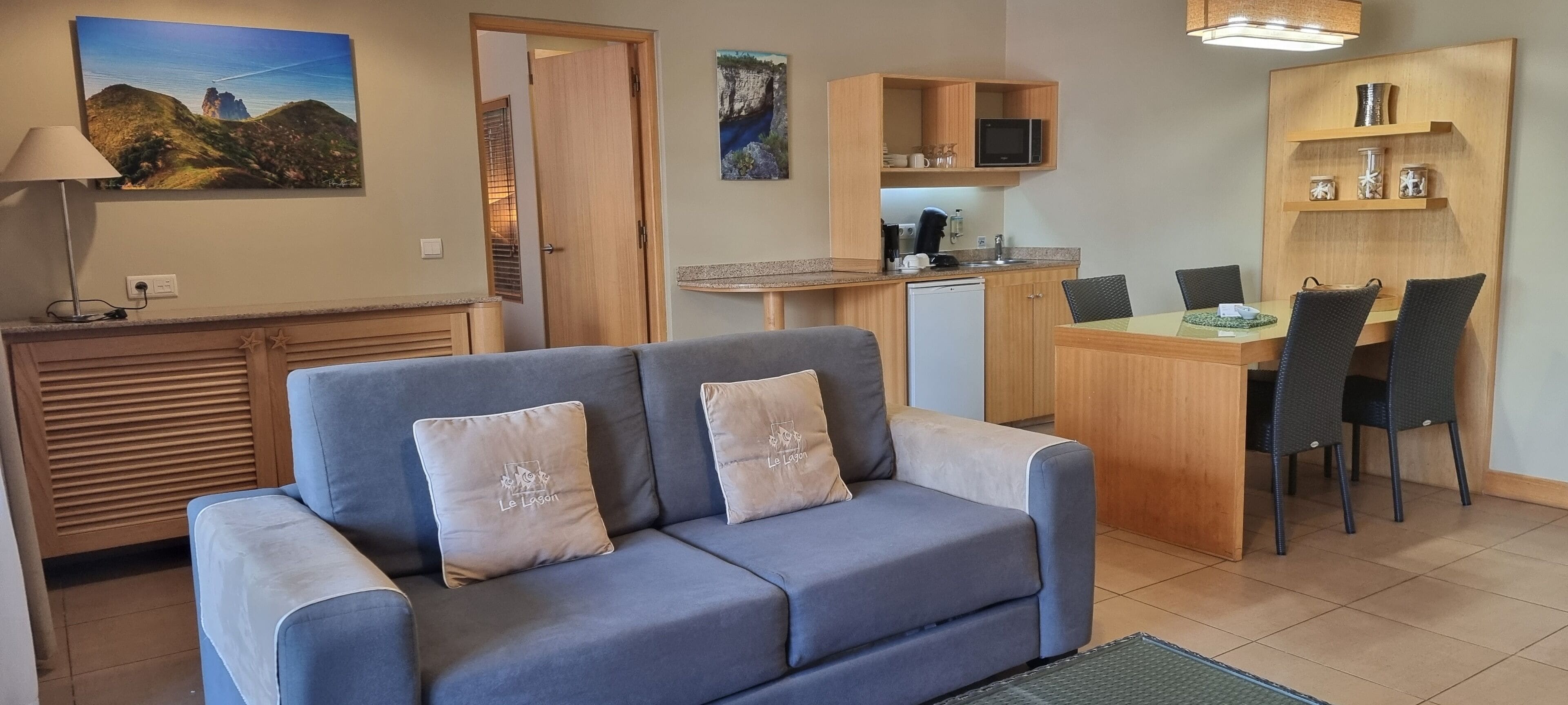 A living room with a sofa, a dining table with chairs, a kitchen area, and a door leading to another room.