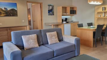 A living room with a sofa, a dining table with chairs, a kitchen area, and a door leading to another room.
