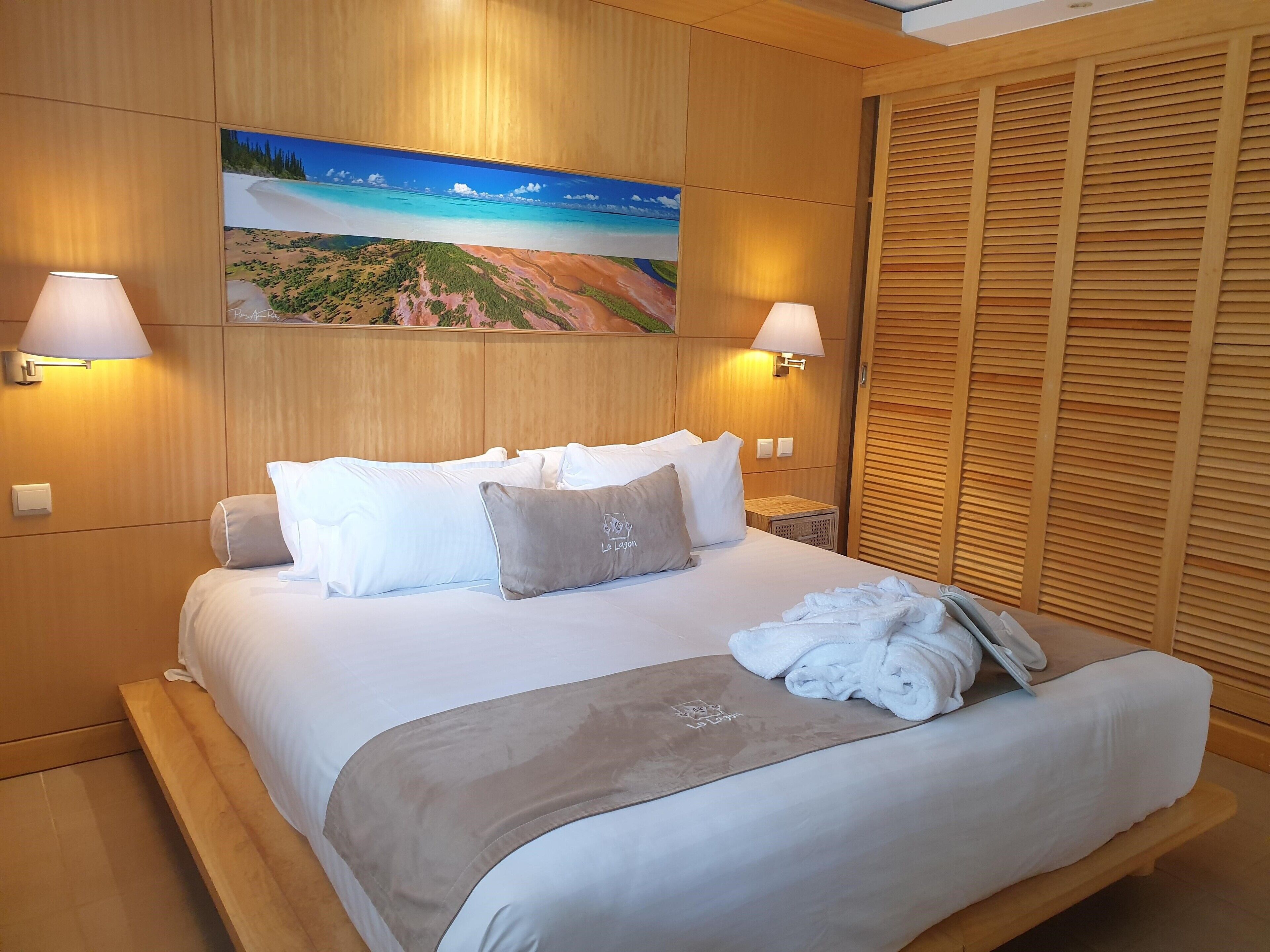 Una cama bien hecha con ropa de cama blanca y gris, una toalla y una colcha. Dos lámparas fijadas a la pared a cada lado de la cama. Un cuadro de una escena de playa está sobre la cama.