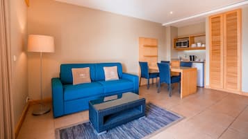 A modern living room with a blue sofa, a coffee table, and a dining area with chairs and a small kitchen in the background.
