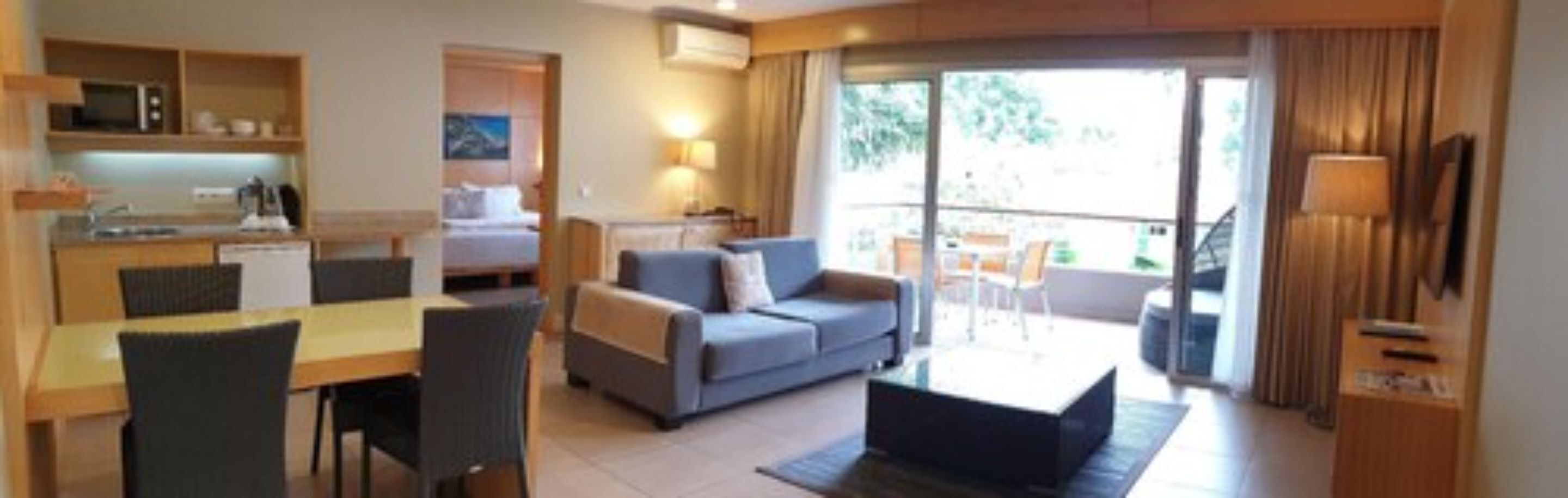 A modern hotel room with a kitchenette, a sofa, a dining area, and a balcony with a view.