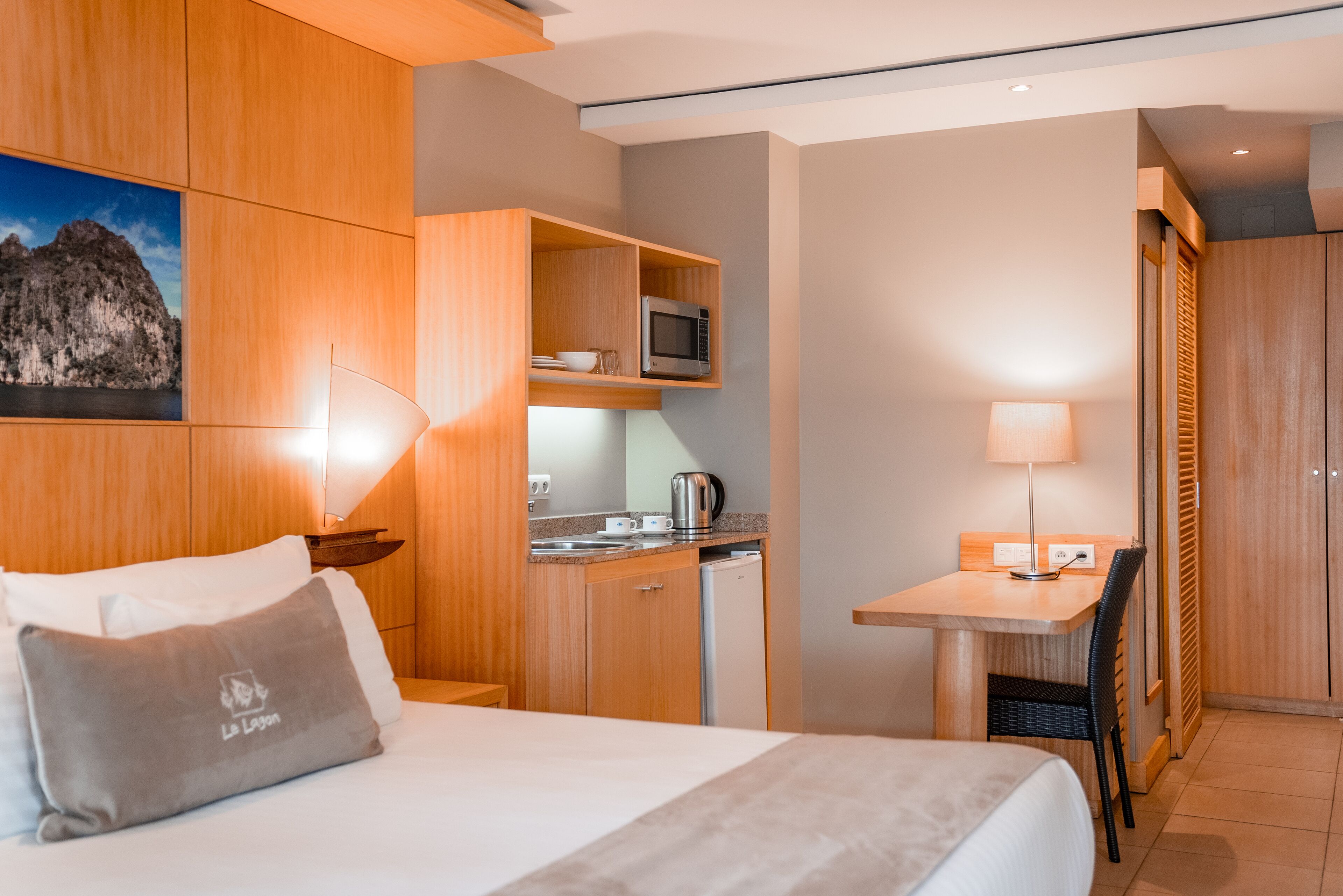 A compact hotel room with a bed, a small kitchenette, a desk, and a dining area.