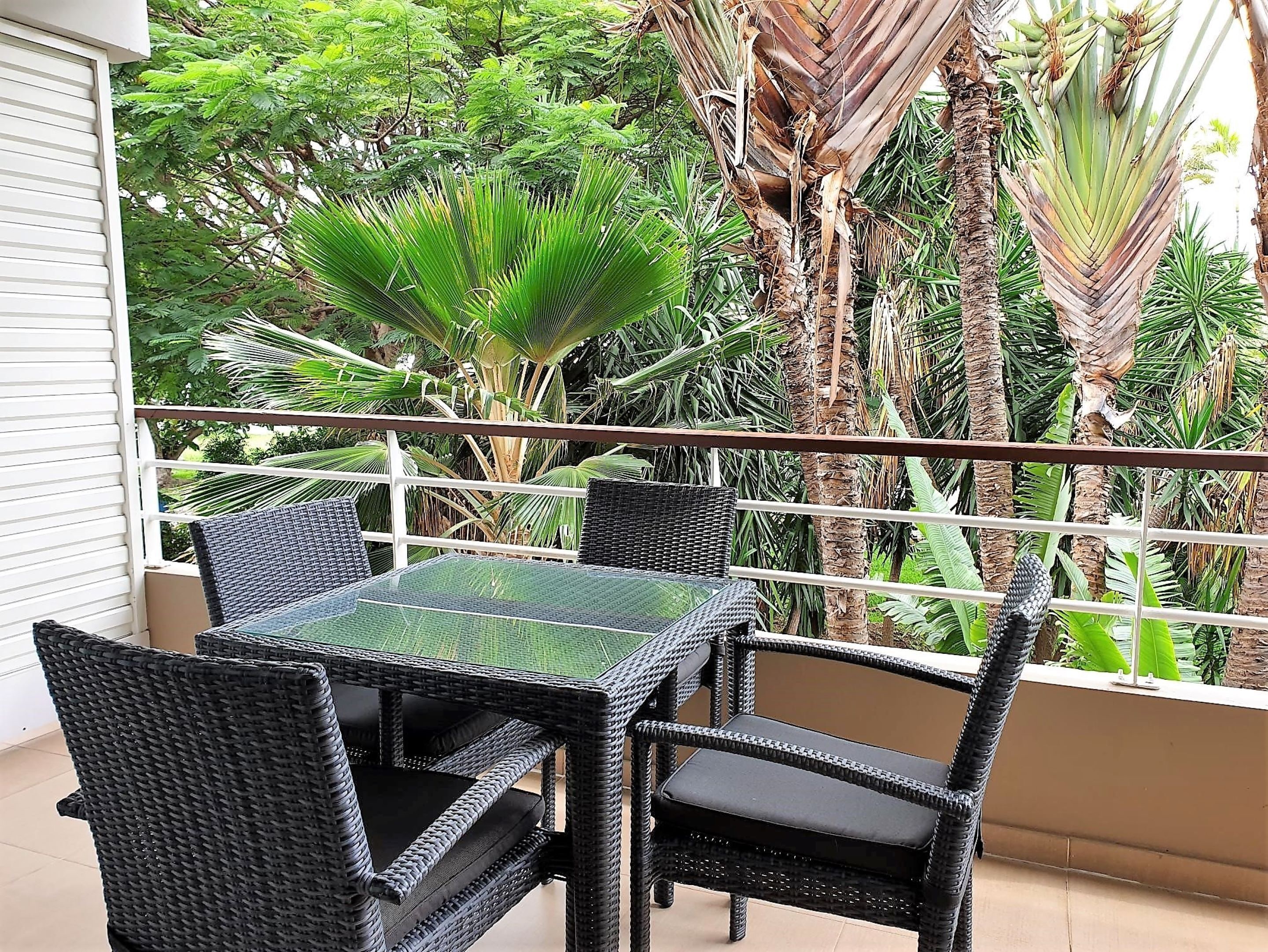 A balcony with a glass table and wicker chairs, surrounded by lush tropical plants.