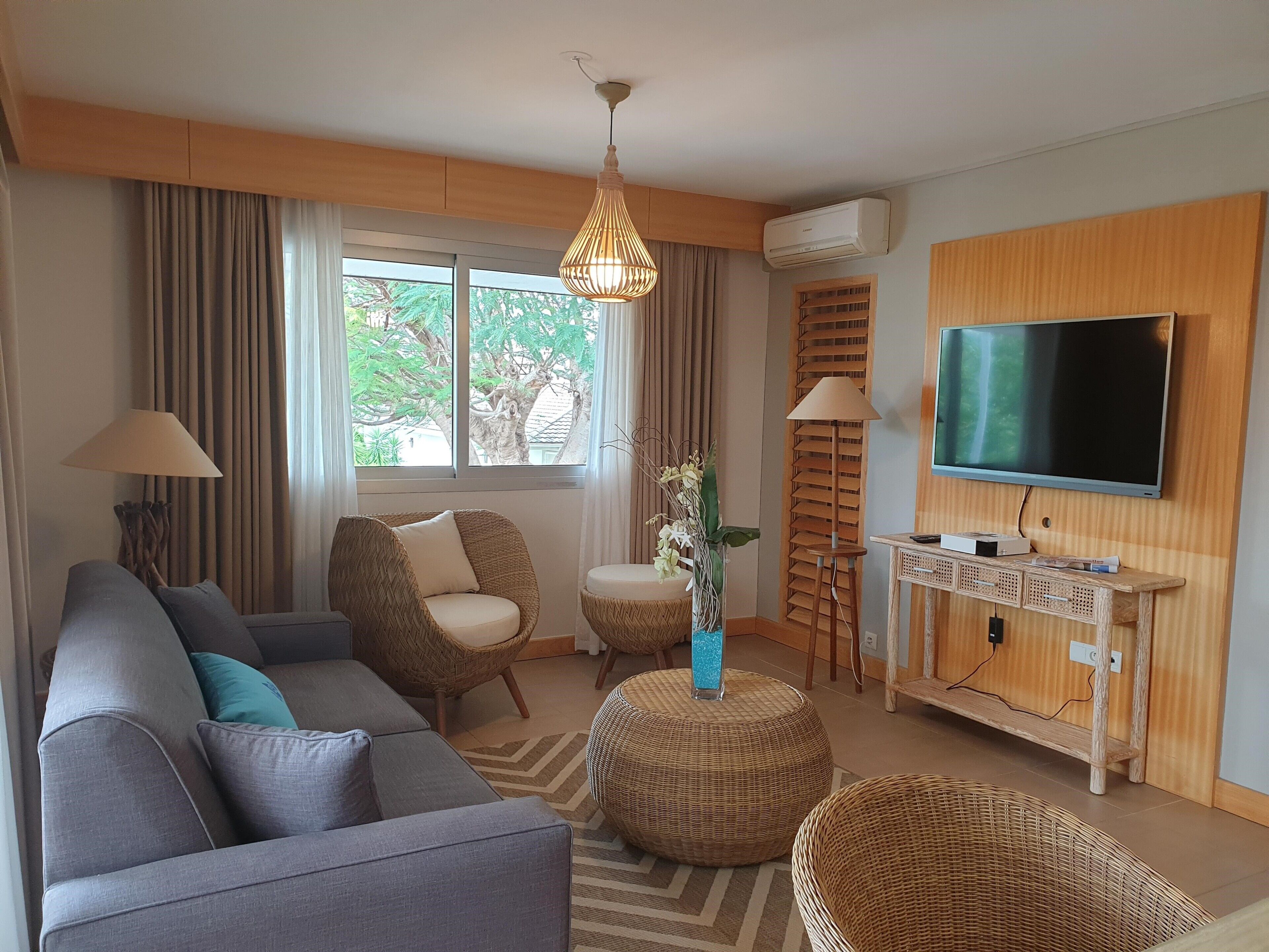 A modern living room with a grey sofa, wicker chairs, a television, and a large window with curtains.