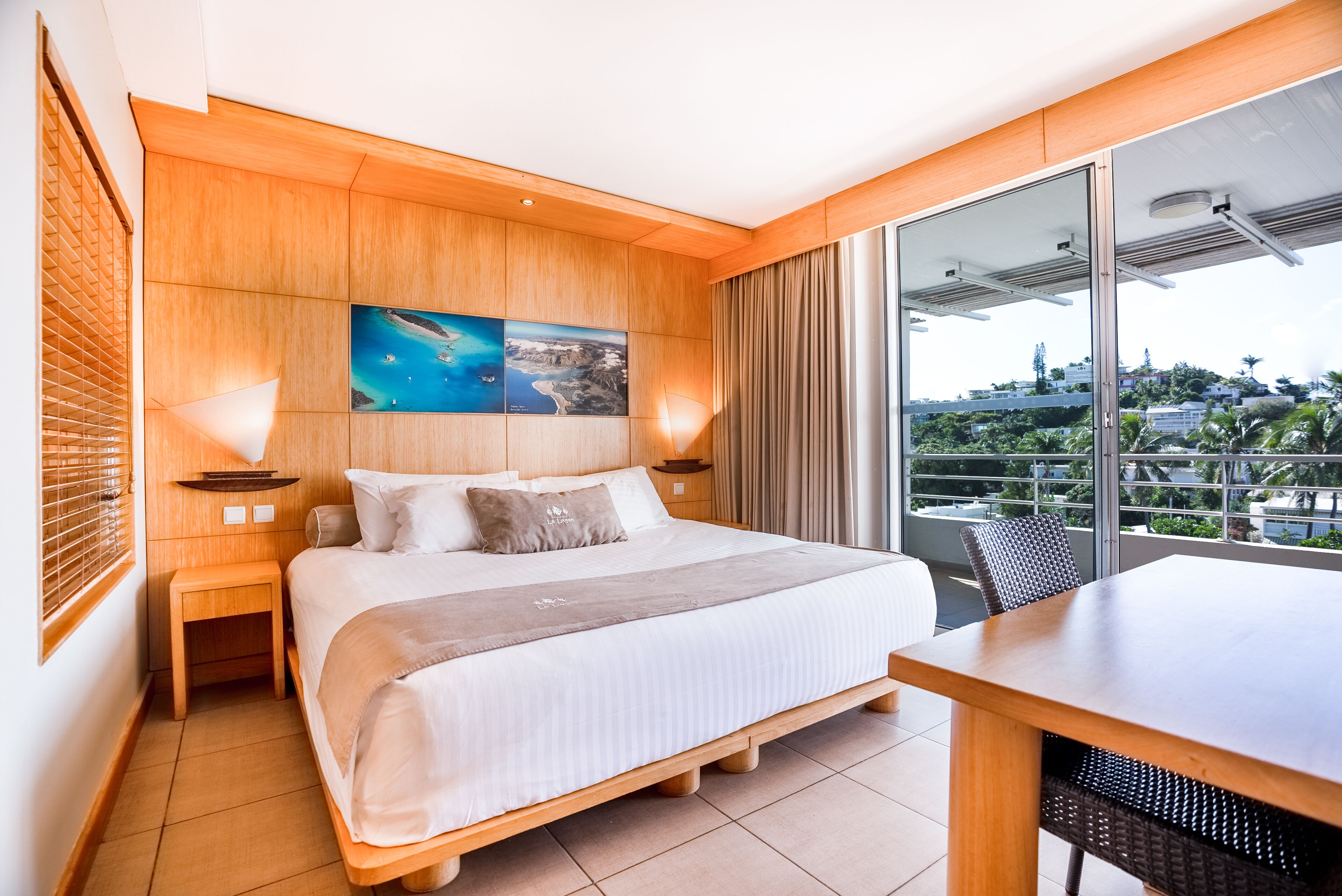 A hotel room with a large bed, wooden headboard, a painting above the bed, a balcony with a view, and a dining area.