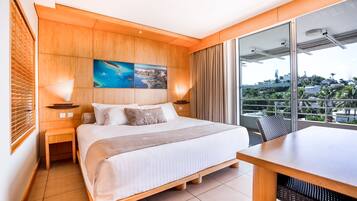 A hotel room with a large bed, wooden headboard, a painting above the bed, a balcony with a view, and a dining area.
