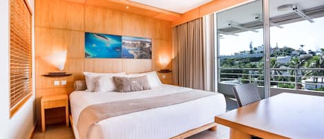A hotel room with a large bed, wooden headboard, a painting above the bed, a balcony with a view, and a dining area.