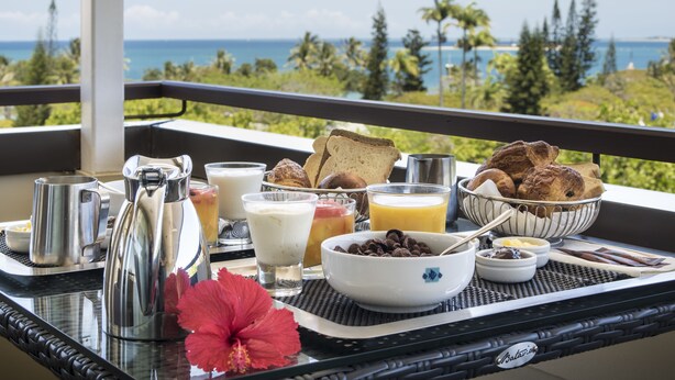 Un plateau de petit-déjeuner avec du café, du lait, du jus de fruits et des viennoiseries, posé sur un balcon offrant une vue panoramique.