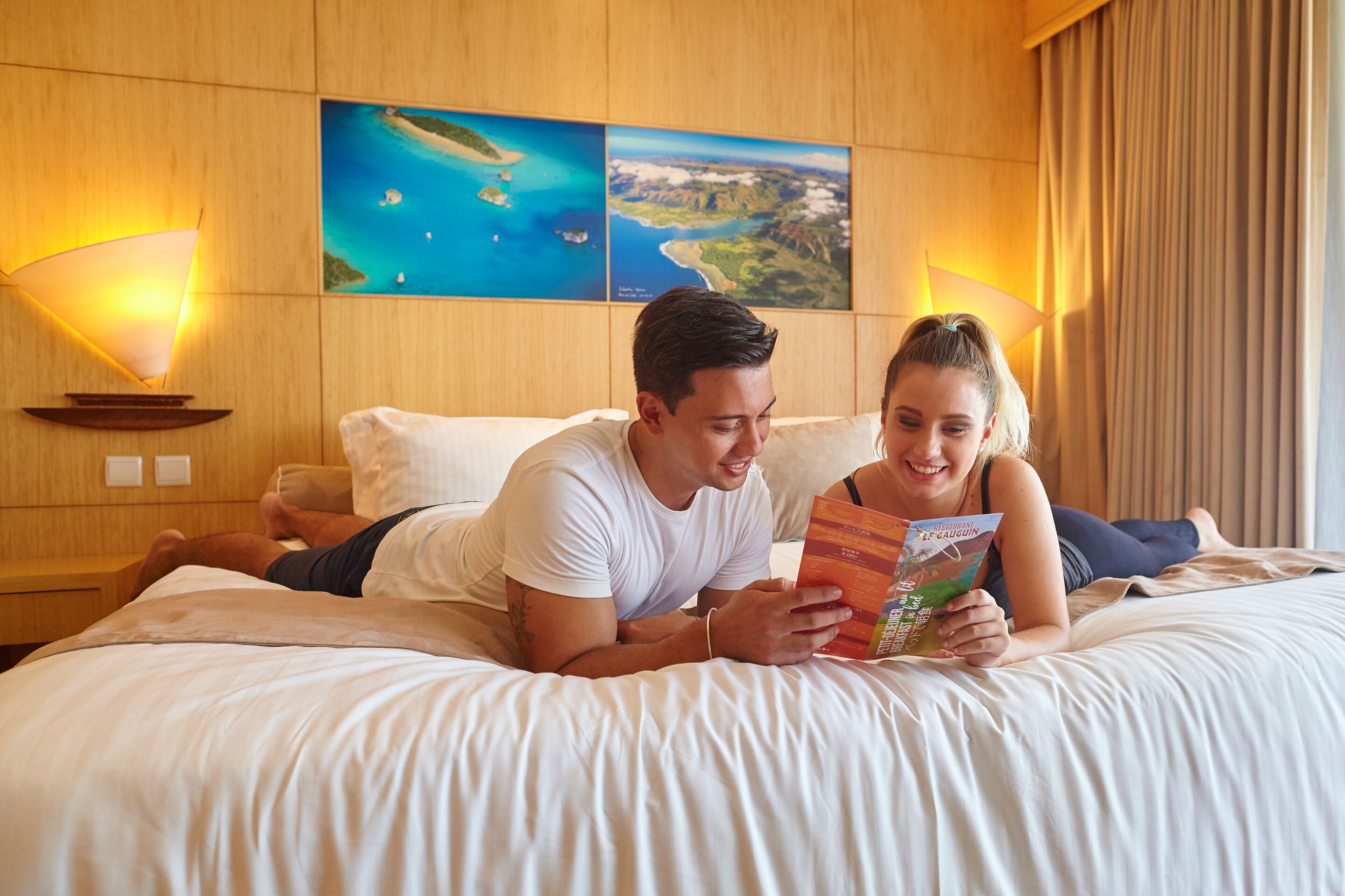 A couple lying on a bed in a hotel room with a view of a coastal landscape and two wall-mounted lamps.