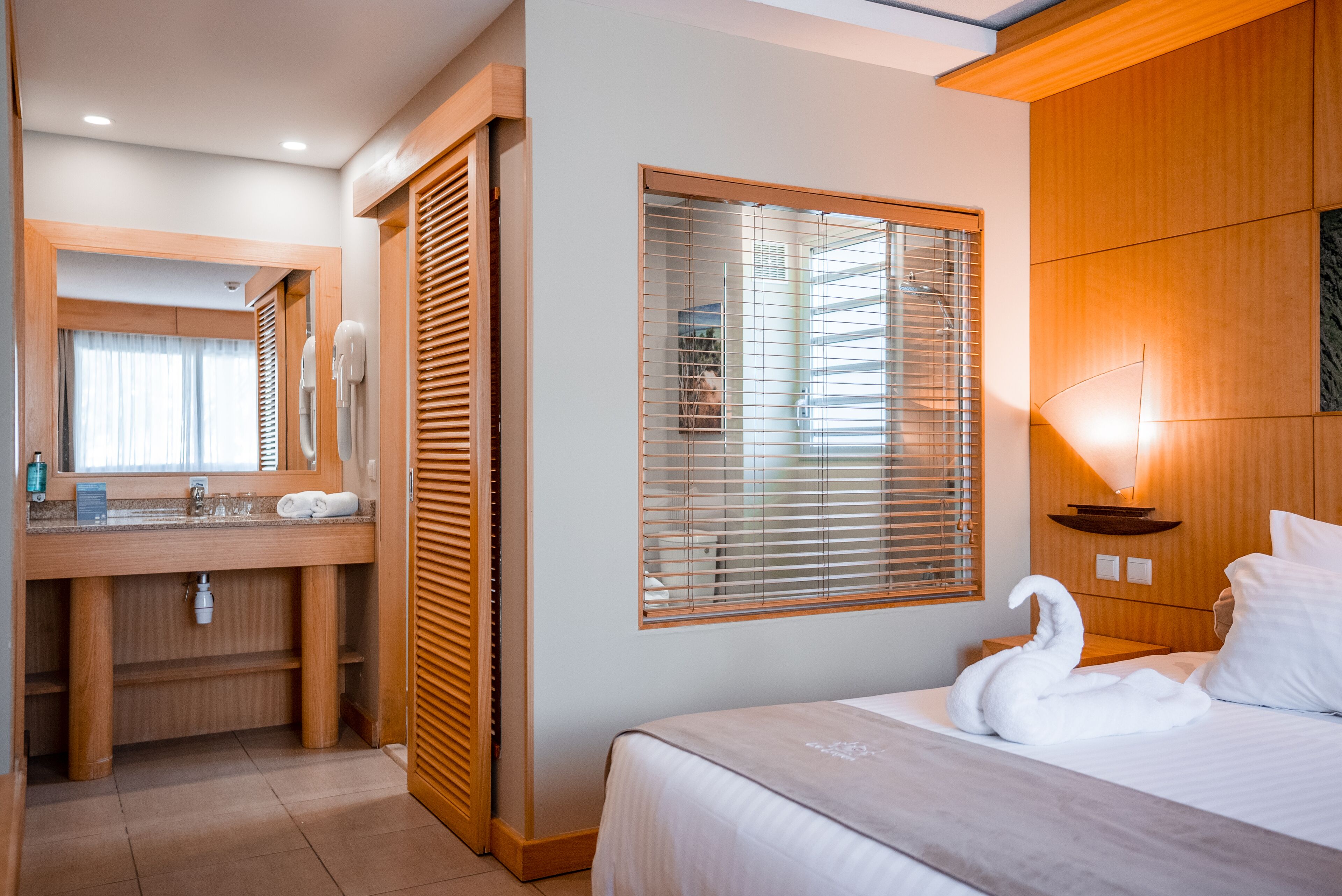 A hotel room with a wooden vanity, a large window with blinds, a bed with a swan-shaped towel, and a nightstand with a lamp.