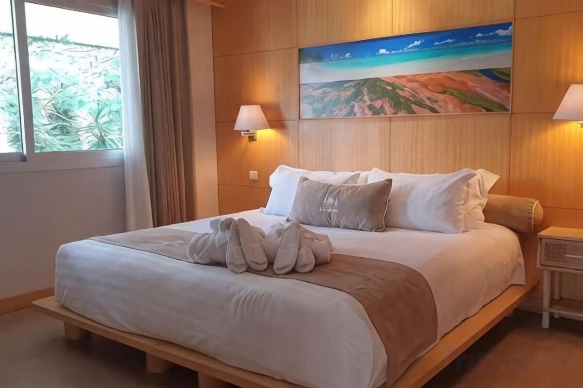 A hotel room with a bed, pillows, a painting on the wall, and a lamp.