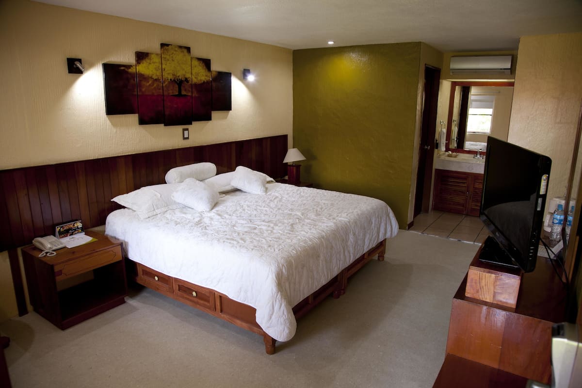A hotel room with a large bed, wooden headboard, a bedside table with a phone, a wall-mounted TV, and a painting above the bed.