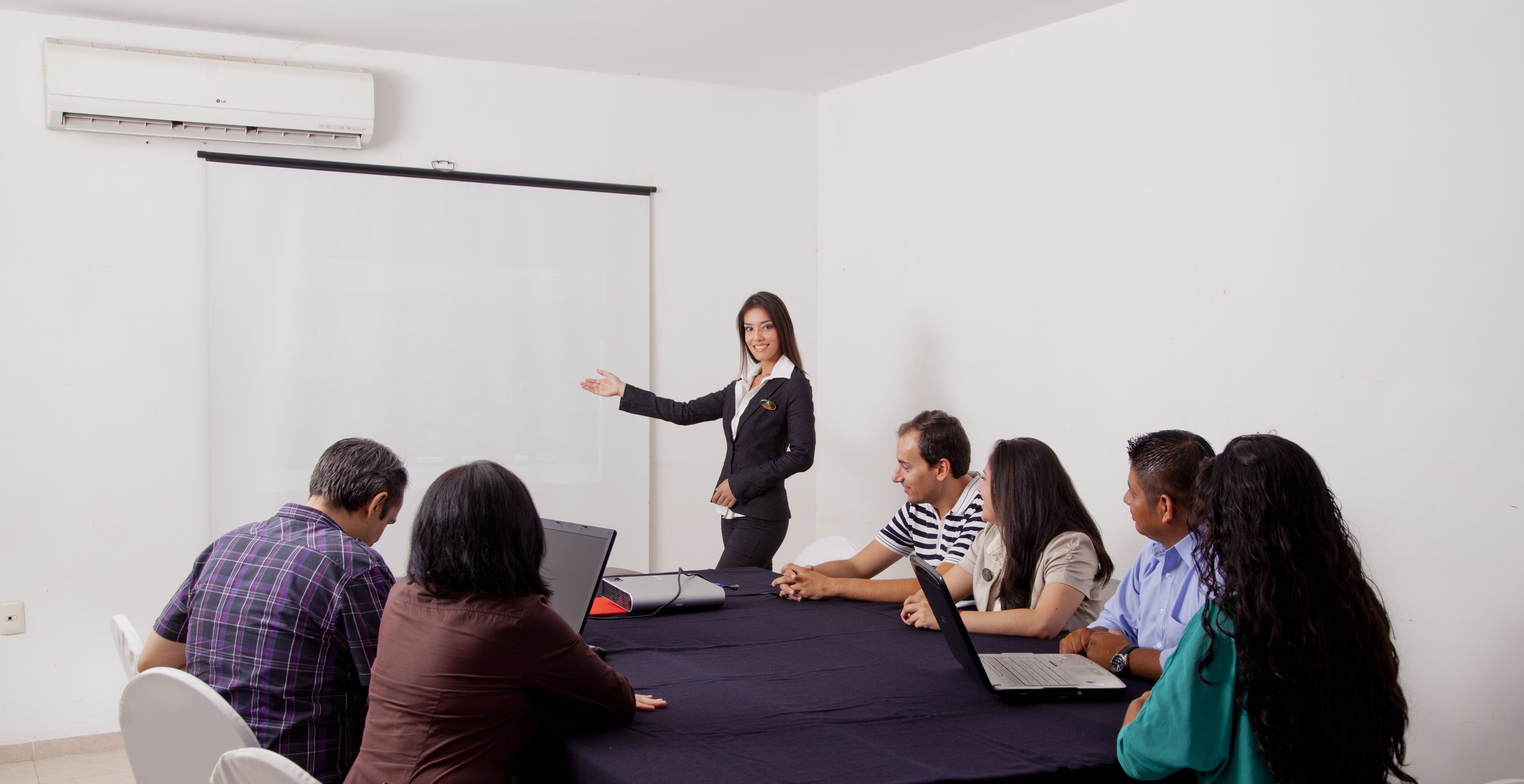 A meeting room with a projector screen, a table with laptops, and a person presenting.