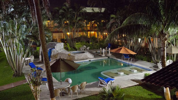 A nighttime view of a resort pool area with lounge chairs, umbrellas, and a stone hot tub. - Hotel Los Andes (Coatzacoalcos)