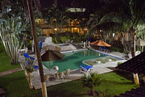 Vista nocturna de un área de piscina en un complejo turístico, con sillones de descanso, sombrillas y un jacuzzi de piedra.