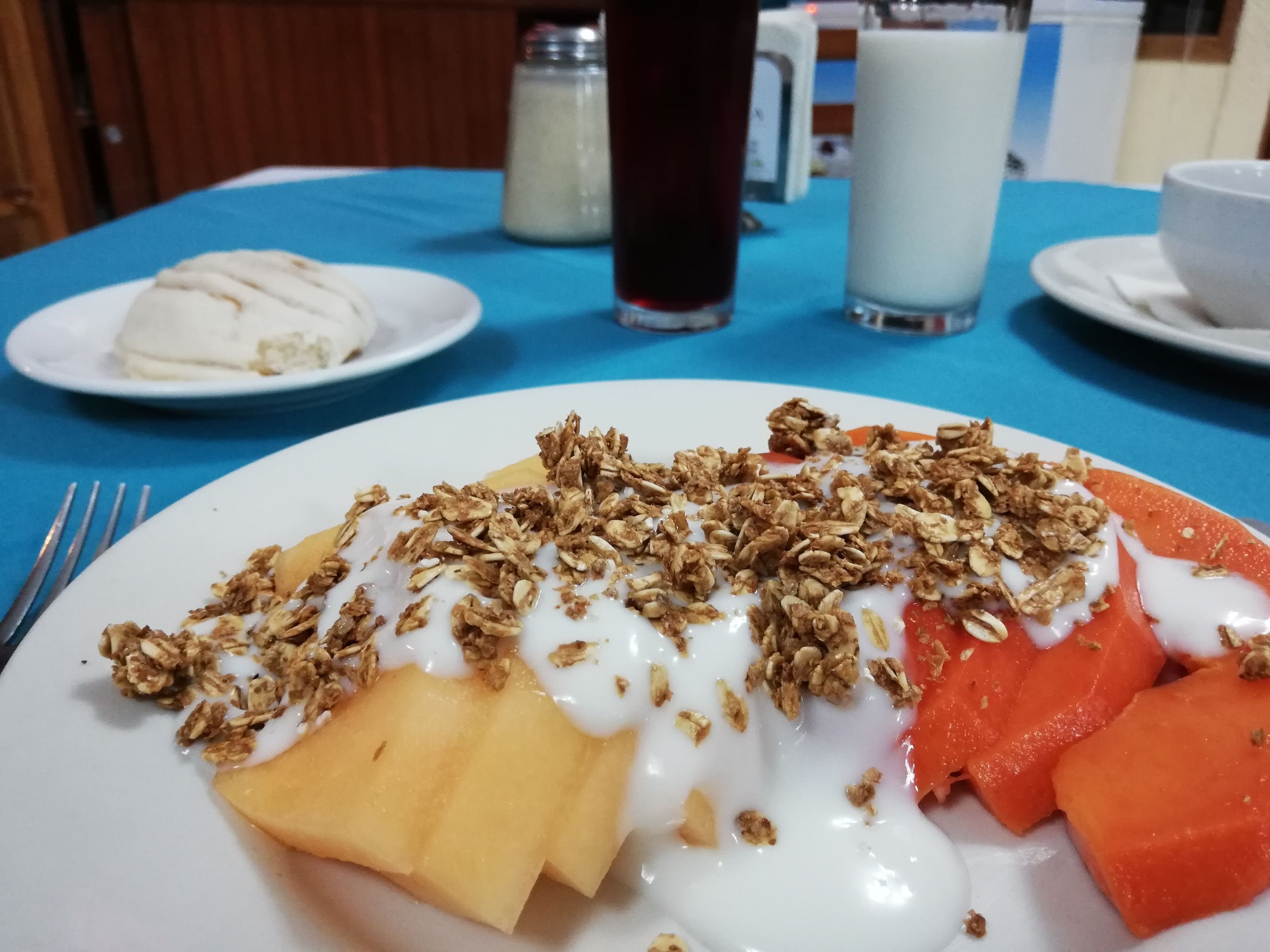 En tallrik med fruktsallad, granola och yoghurt, ett glas med mörk vätska och en kopp mjölk står på ett bord med en blå duk.