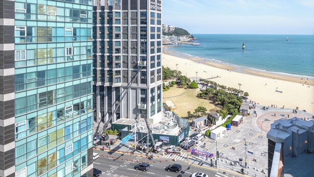 A high-rise building with a modern glass facade, a beach in the background, and a clear sky. - Sunset Hotel (Busan)
