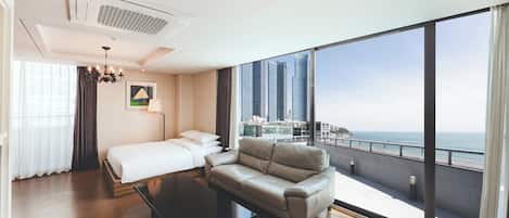 A modern hotel room with a bed, a sofa, a coffee table, and a large window offering a city view.