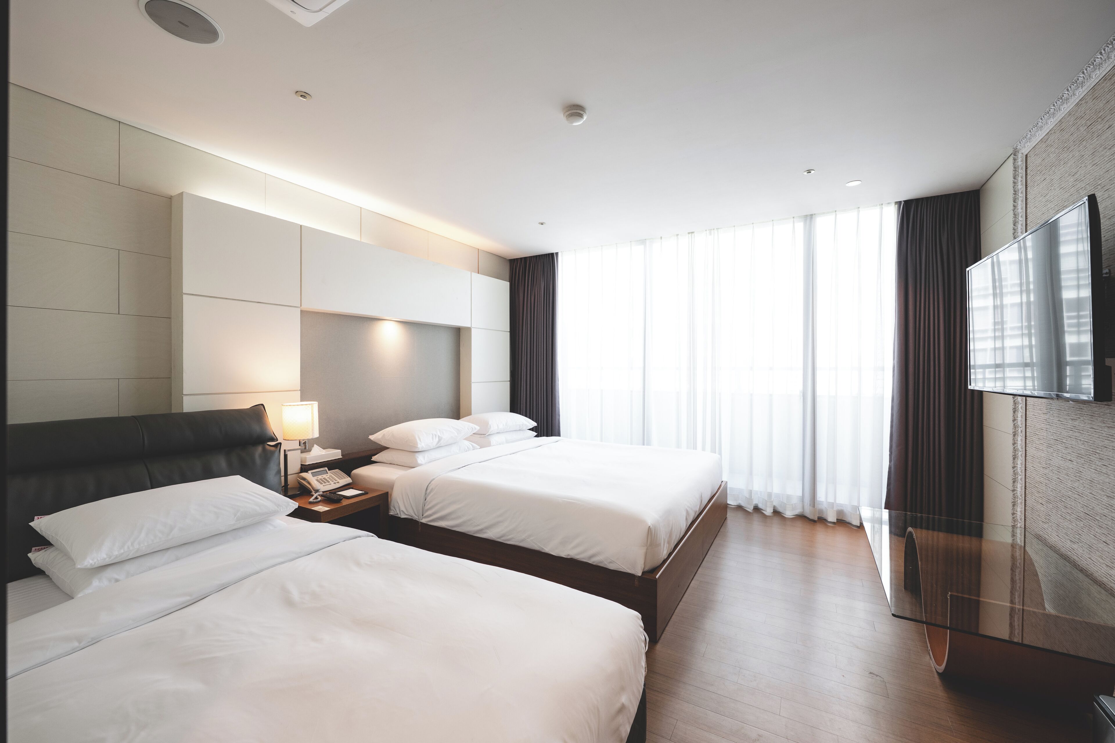 Habitación de hotel con dos camas, una mesita de noche con lámpara, un televisor de pantalla plana montado en la pared y ventanas amplias con cortinas.