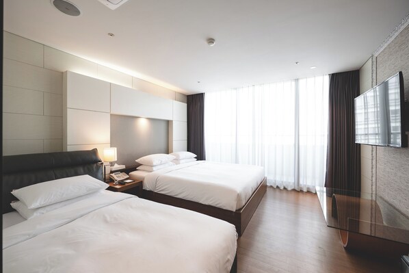 A hotel room with two beds, a bedside table with a lamp, a flat-screen TV mounted on the wall, and large windows with curtains.