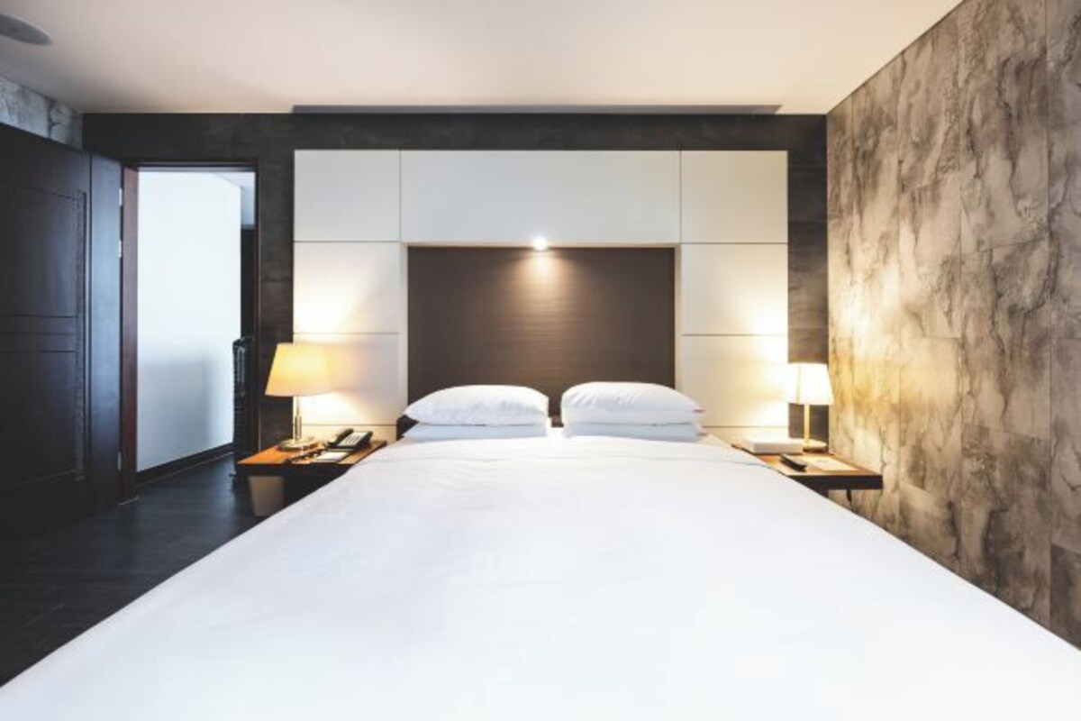 A modern hotel room with a large bed, two bedside tables with lamps, a dark headboard, and a marble accent wall.
