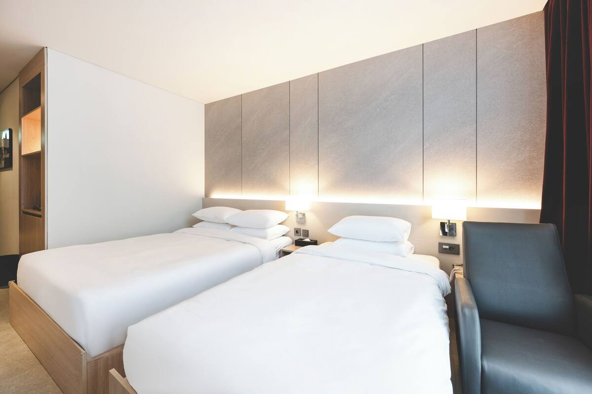 A hotel room with two single beds, a wooden wardrobe, a dark armchair, and a wall-mounted TV.