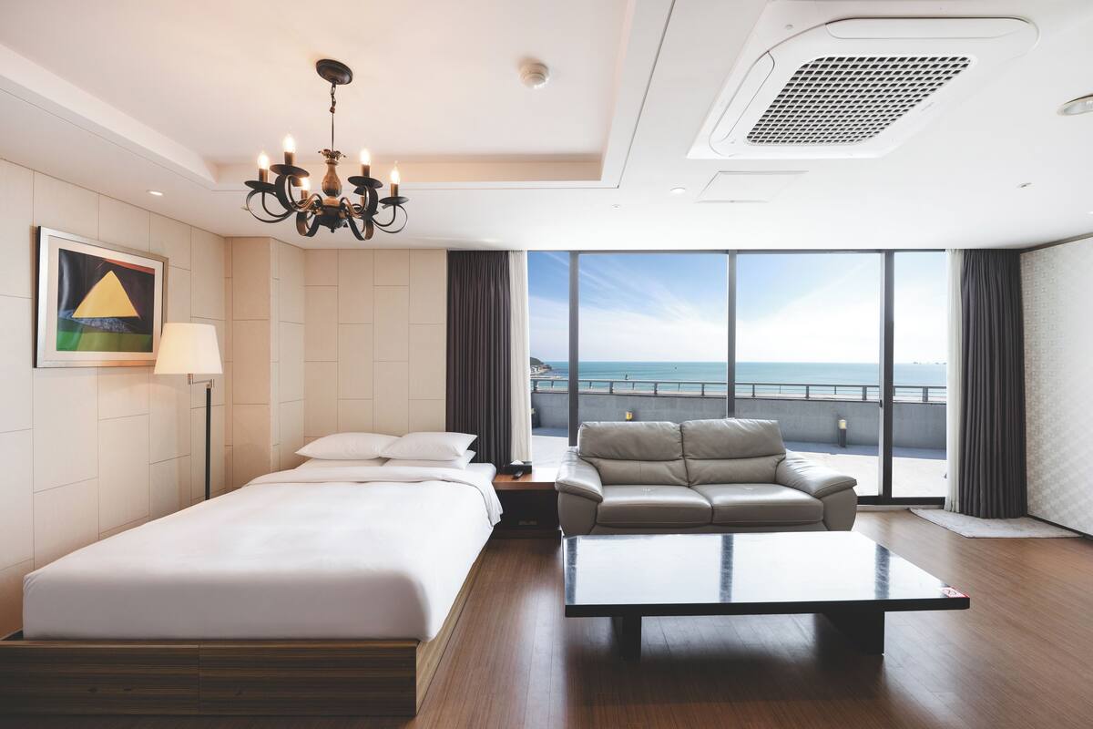 A hotel room with a bed, a sofa, a coffee table, a chandelier, and a large window with a view of the sea.