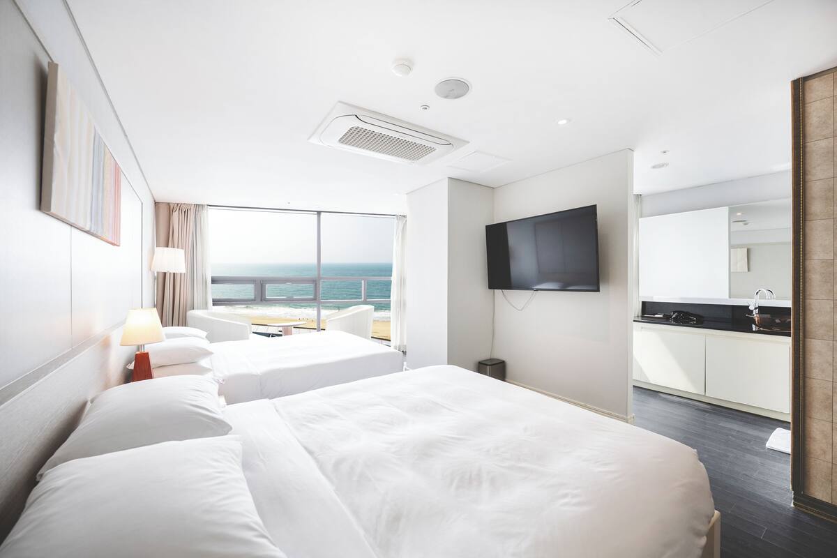 A hotel room with two beds, a TV, a balcony with a view of the sea, and a kitchenette.