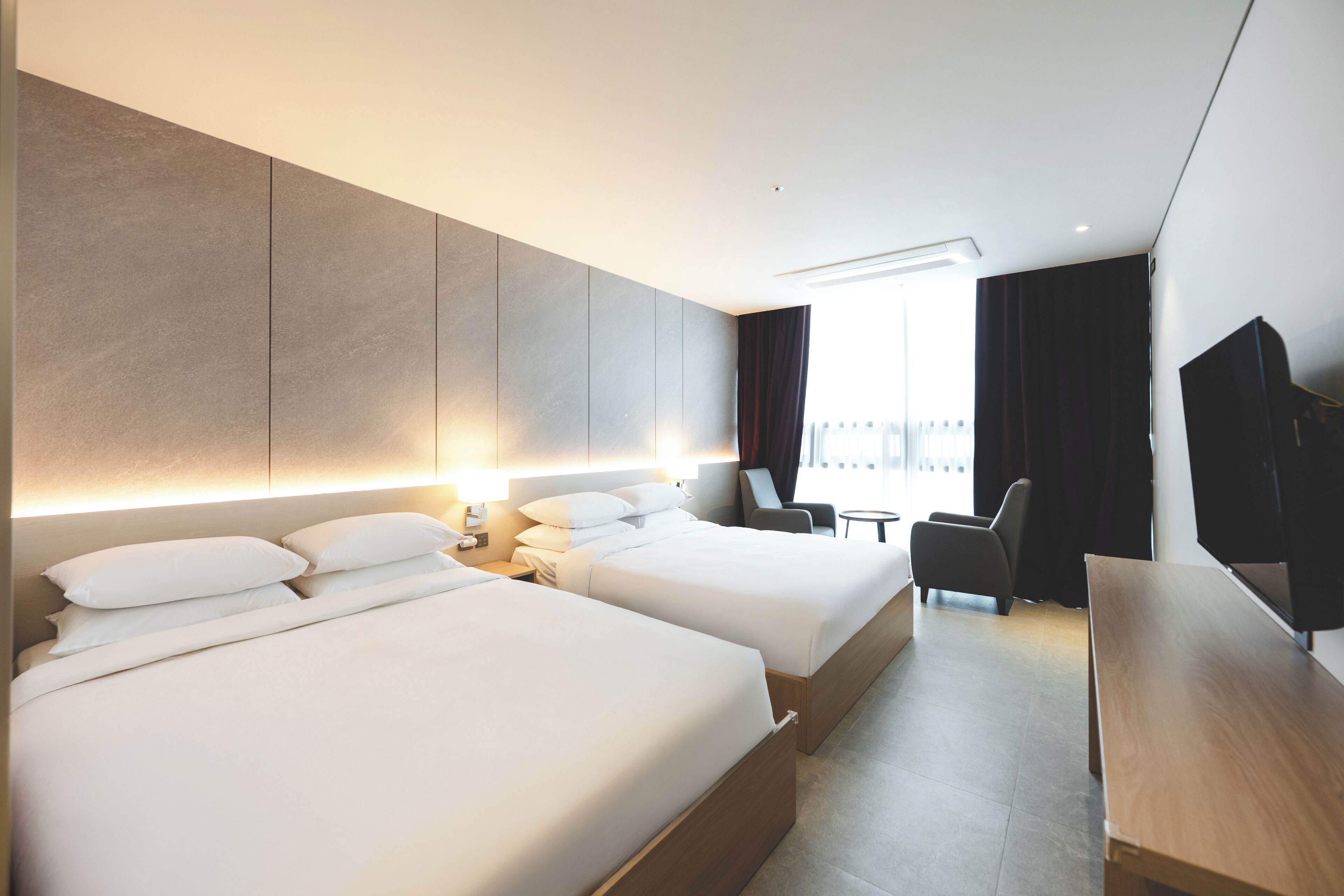 A modern hotel room with two beds, a desk, and a view of the city.