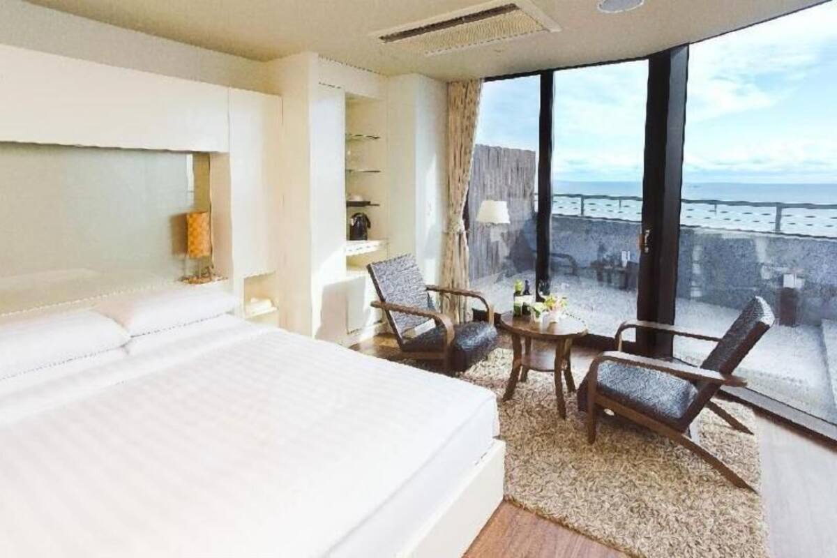 A hotel room with a large bed, two chairs, a small table, and a view of the sea through large windows.