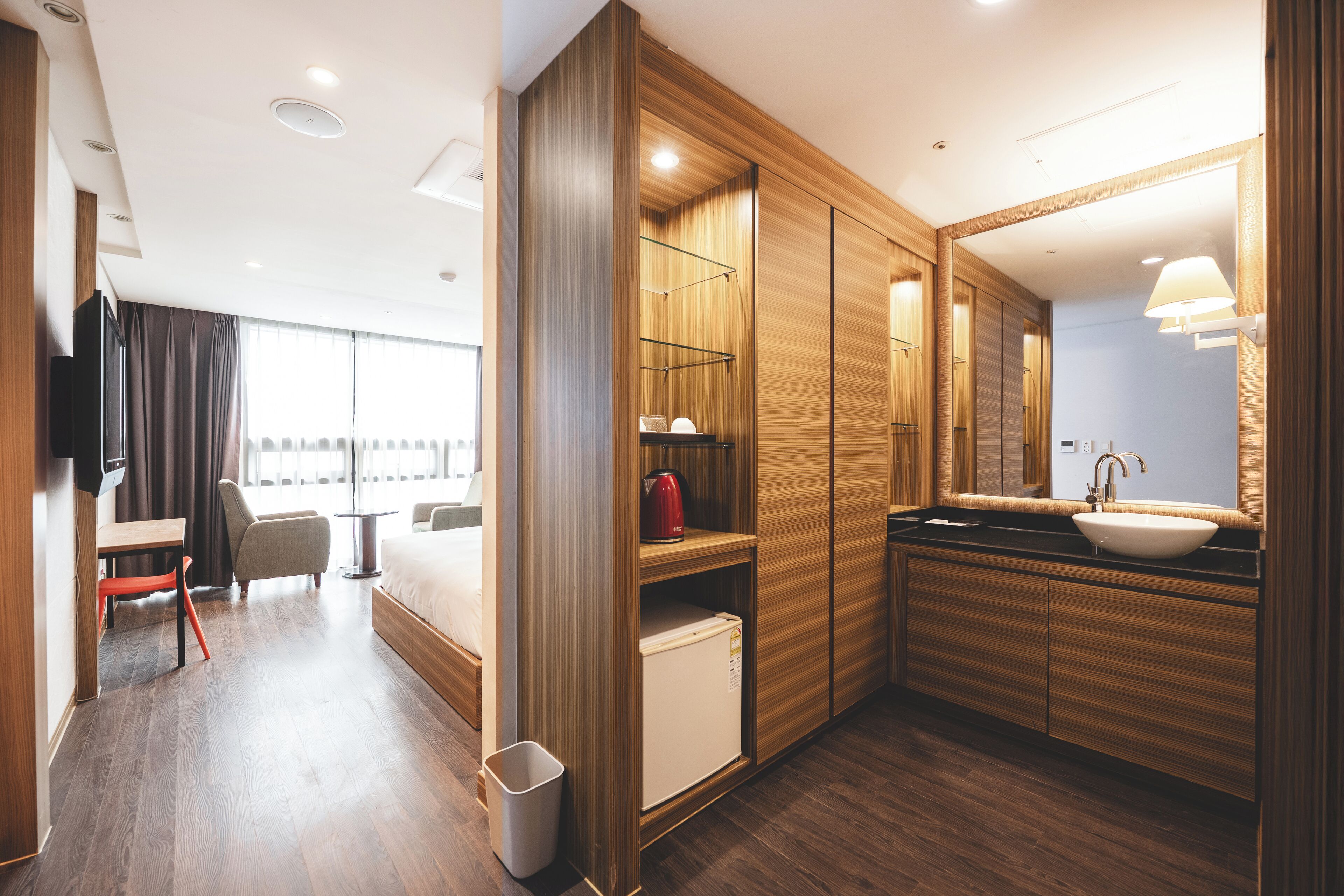 A modern hotel room with a wooden floor, a bed, a desk, a chair, a TV, a wardrobe, a mirror, and a sink.