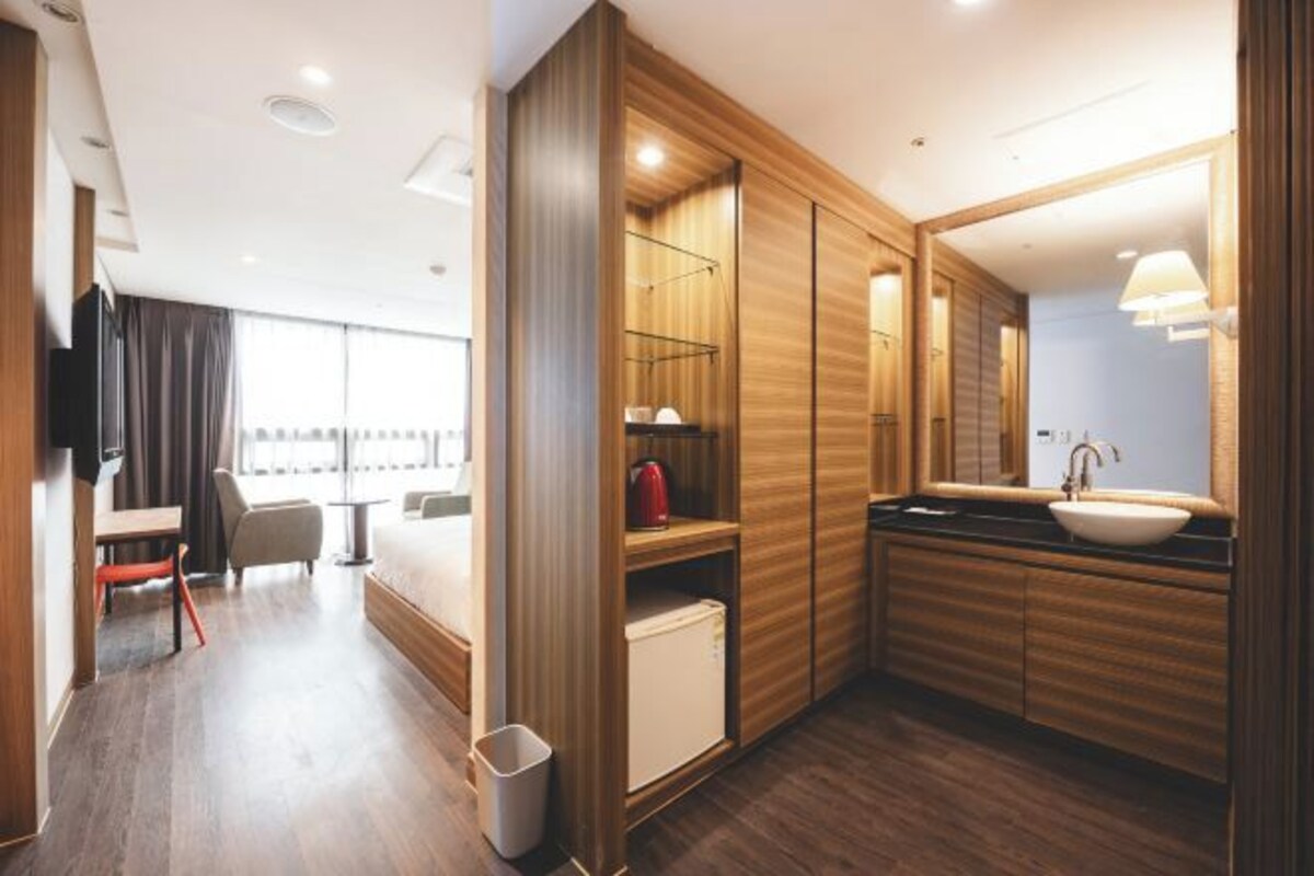 A modern hotel room with a wooden interior, a bed, a desk, a chair, a TV, a large window, a bathroom with a sink and mirror, and a small refrigerator.