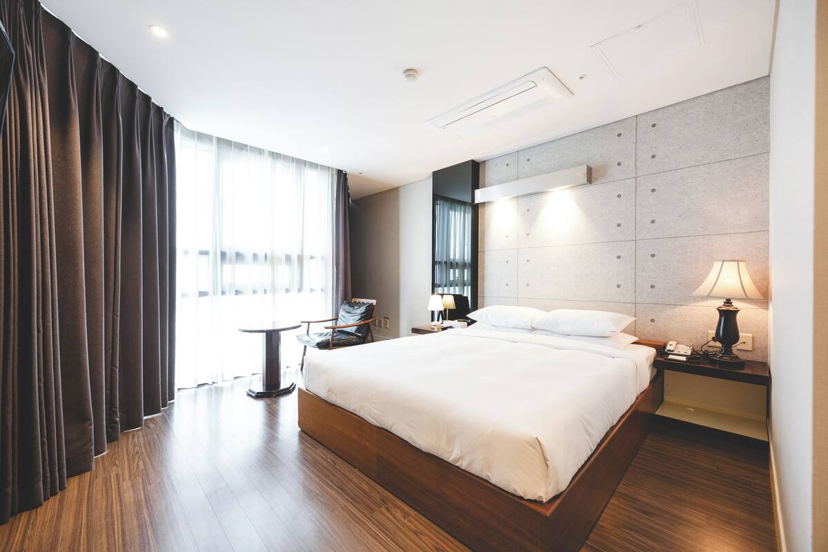 A modern hotel room with a large bed, a bedside table with a lamp, a chair, and a view of the outdoors through large windows.