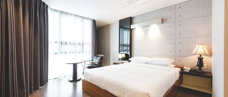 A modern hotel room with a large bed, a bedside table with a lamp, a chair, and a view of the outdoors through large windows.