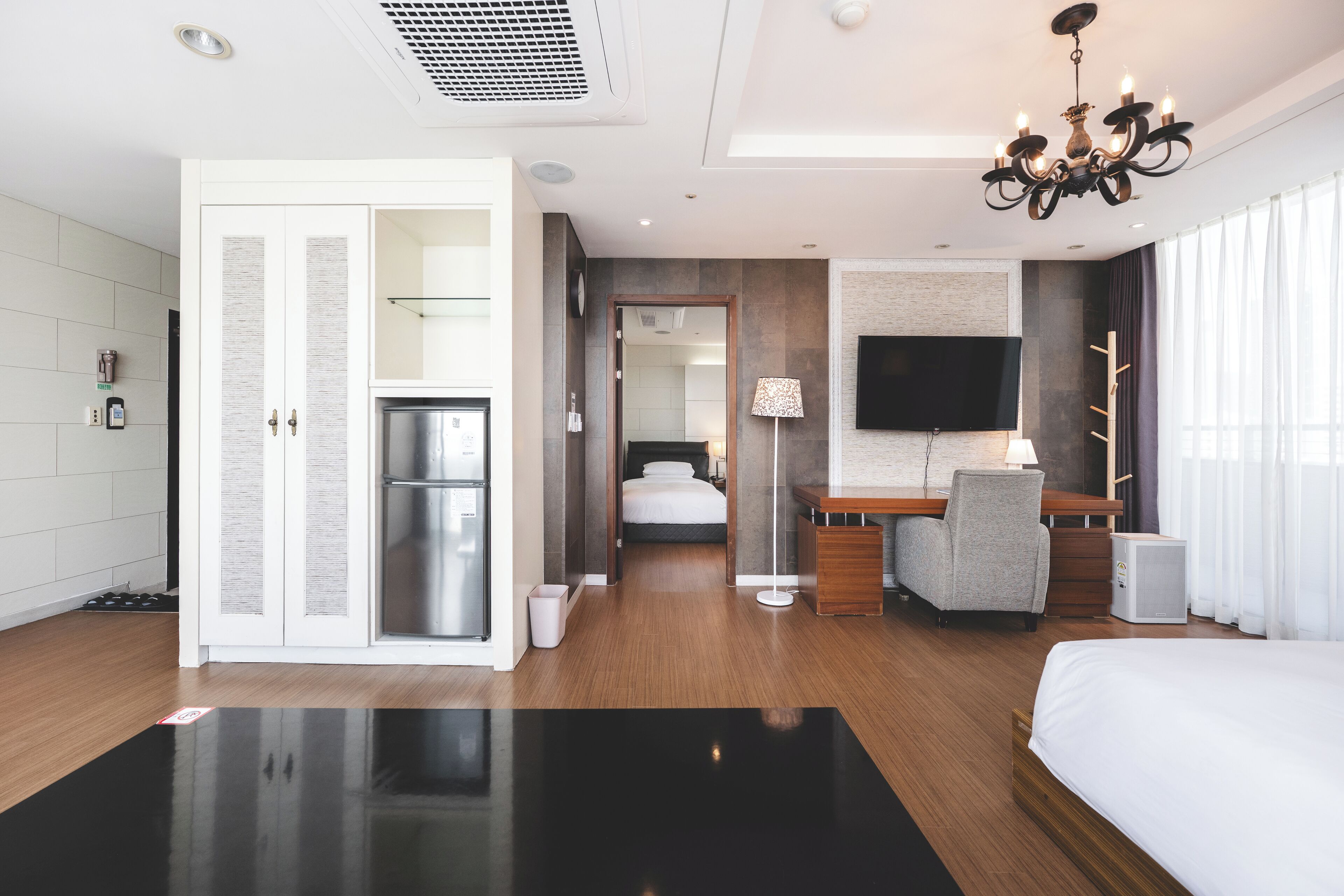 A modern hotel room with a bed, desk, and seating area. It includes a TV, a small fridge, and a bathroom visible through an open door.