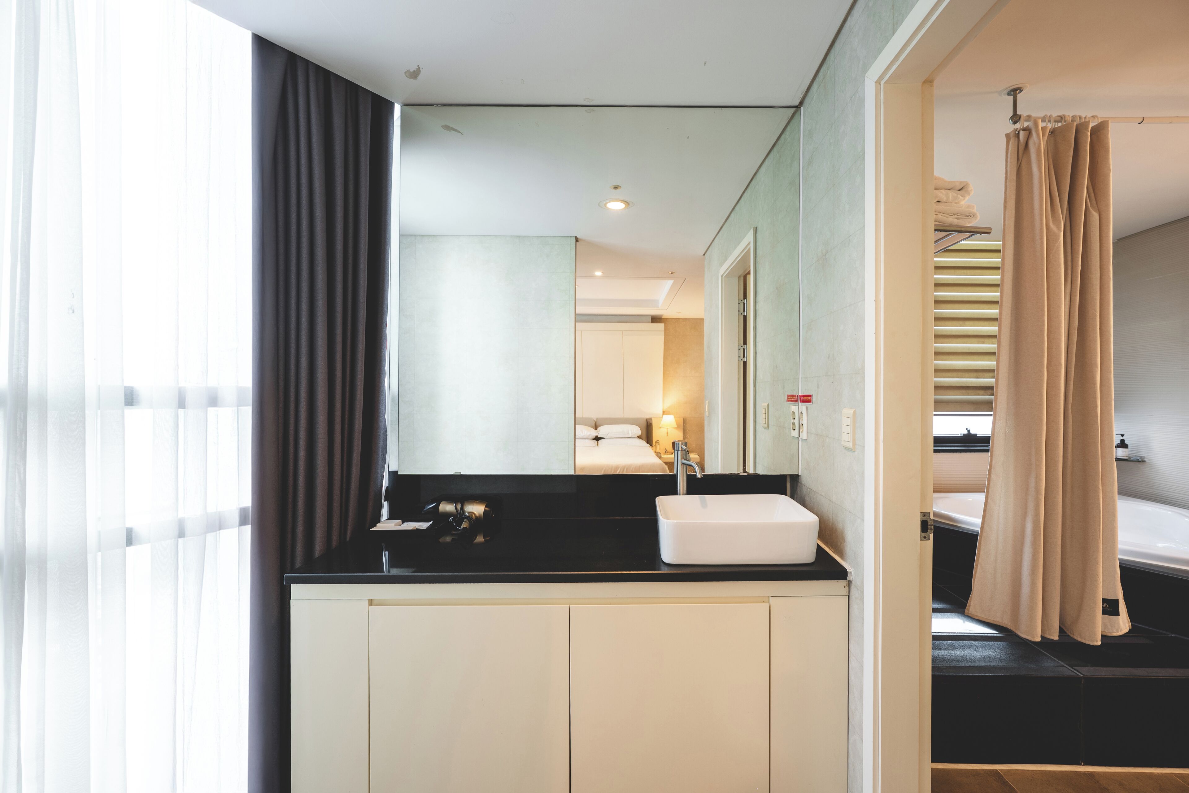 Un baño moderno con encimera negra, lavabo blanco y espejo. Hay una cortina de ducha y se ve una habitación con cama y mesita de noche.