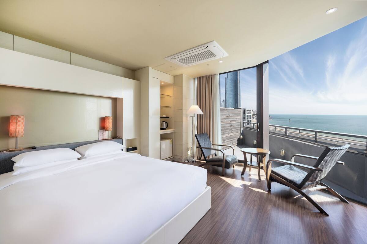 A hotel room with a large bed, a balcony with a view of the sea, a small table with a chair, and a lamp.