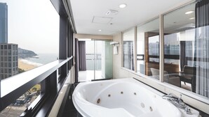 A modern bathroom with a large bathtub, a glass shower enclosure, and a view of the beach and cityscape through large windows. - Sunset Hotel (Busan)