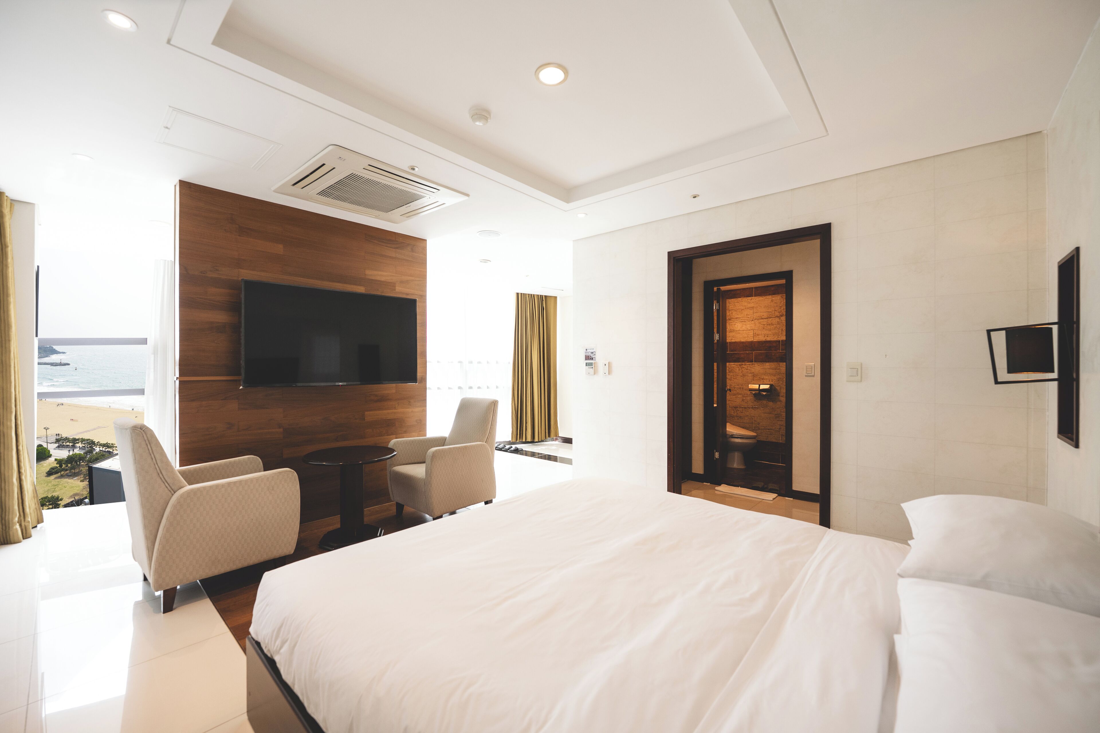Une chambre d’hôtel avec un grand lit, un téléviseur fixé au mur, un coin salon avec des fauteuils et une vue sur la plage.