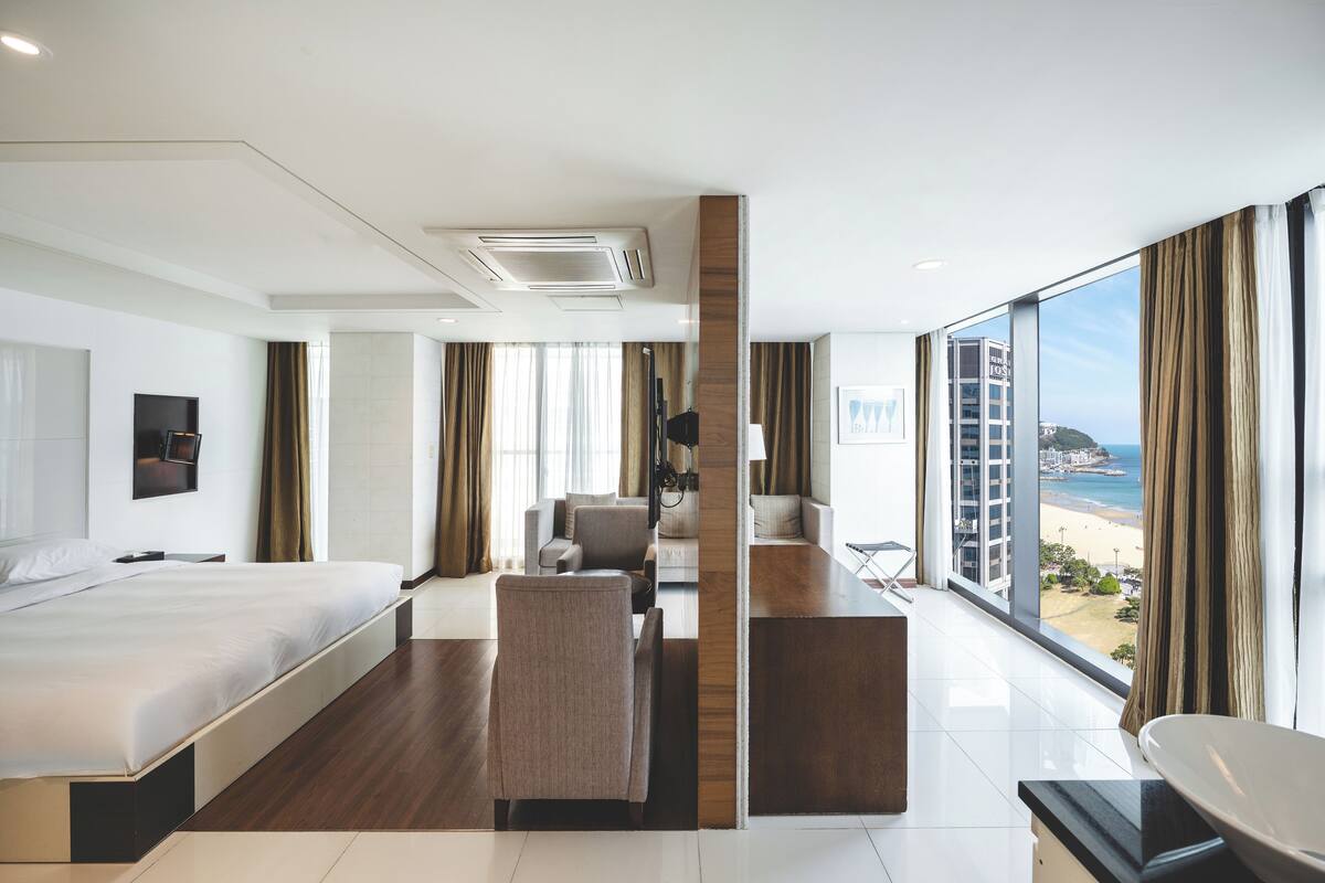 A modern hotel room with a large bed, a seating area with a view of the beach, and a balcony with a cityscape view.