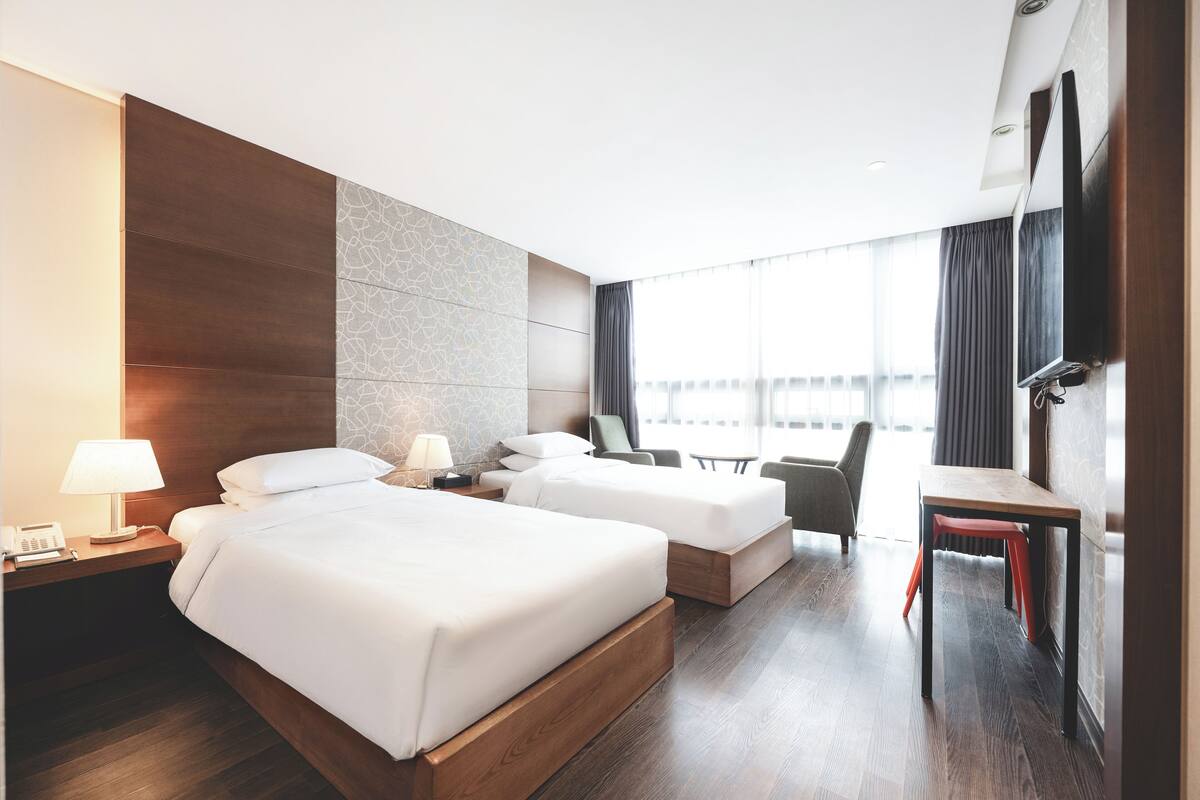 A hotel room with two beds, a desk, a chair, and a large window with curtains.
