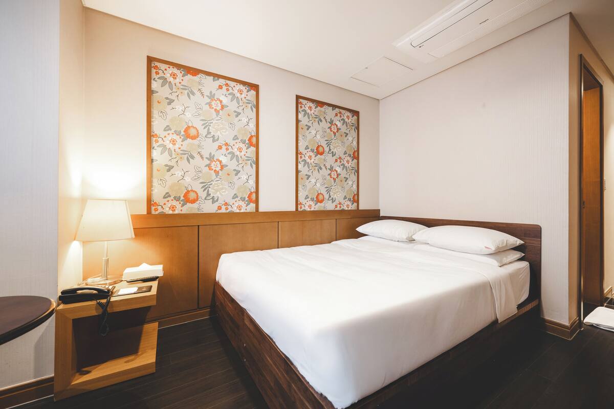 A hotel room with a wooden bed, a bedside table with a lamp, and two floral-patterned wall decorations.
