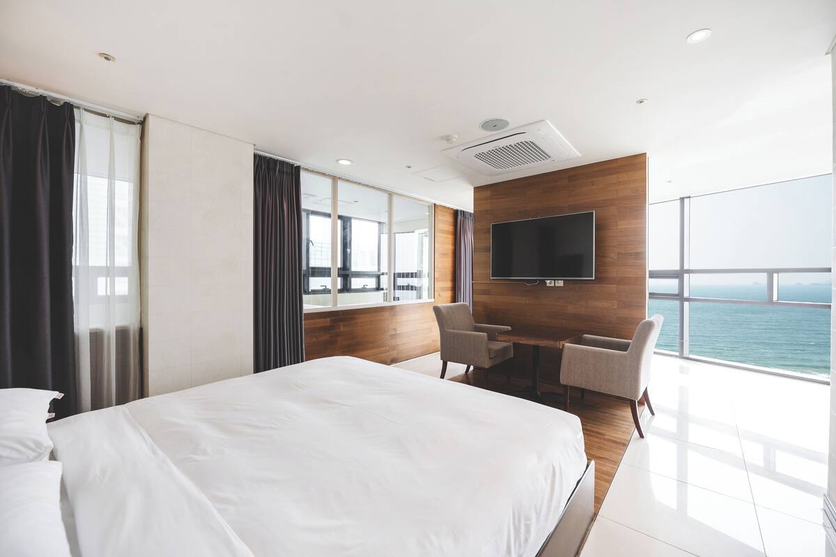 A modern hotel room with a large bed, a flat-screen TV mounted on the wall, a desk with two chairs, and a large window offering a view of the sea.