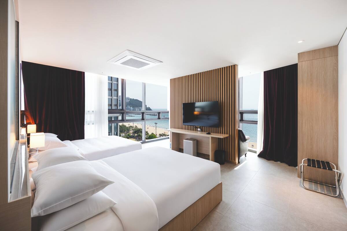 A modern hotel room with two beds, a TV mounted on a wooden panel, and a view of the city and sea.
