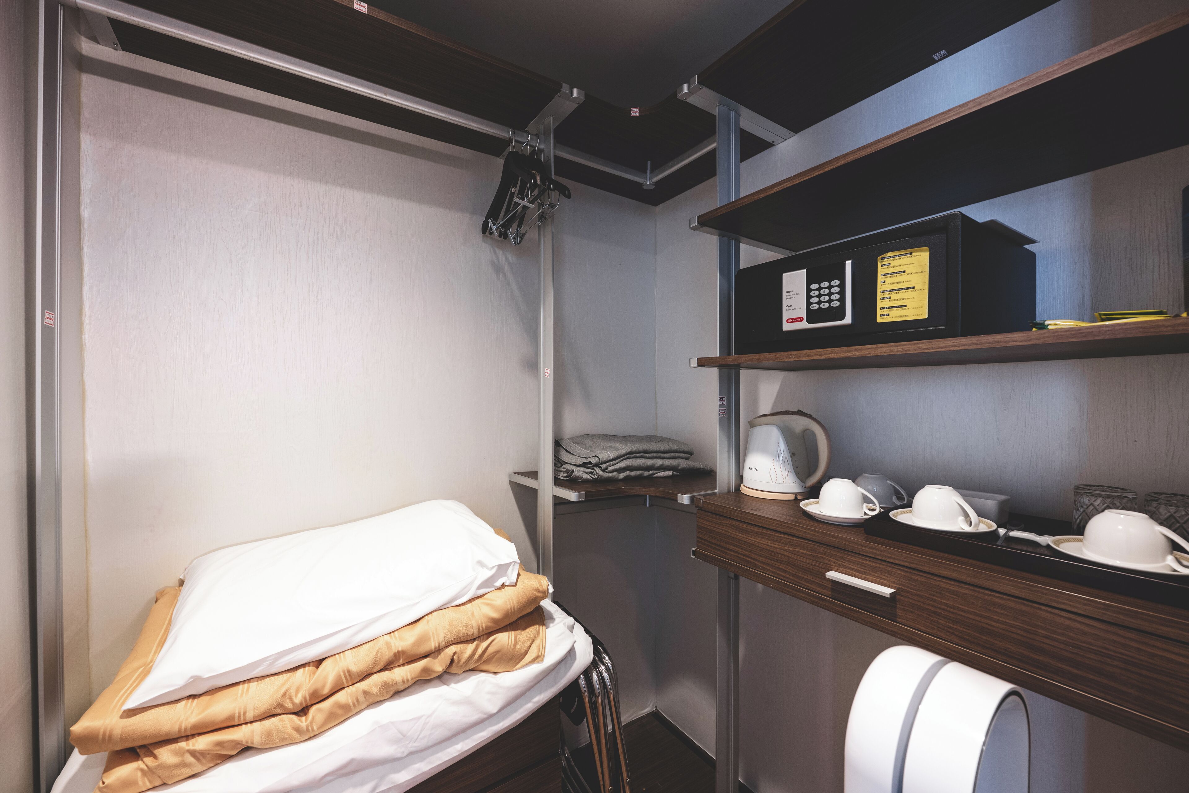 Une chambre d’hôtel compacte comprenant un lit, un petit bureau avec un micro-ondes et une étagère avec des tasses et une bouilloire.